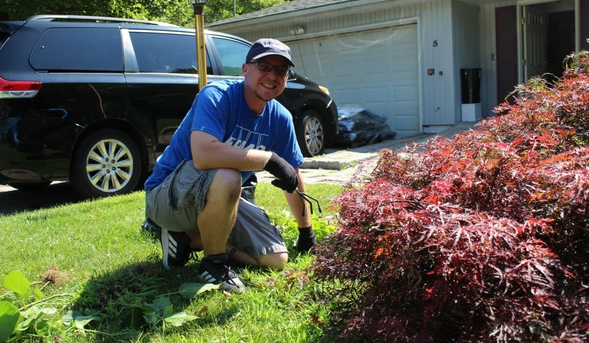Man in a blue shirt kneels in a yard while gardening.