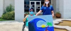 Man seated in a wheelchair and standing woman in a blue shirt hold a large painting of a tropical island scene