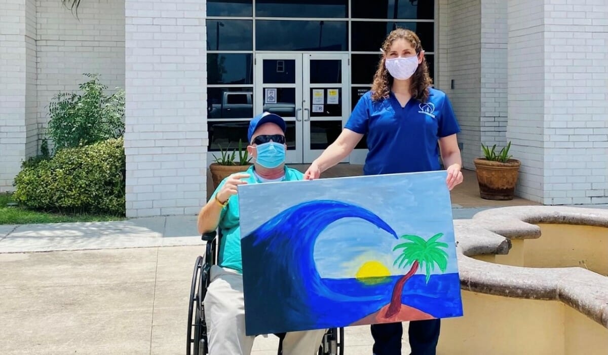 Man seated in a wheelchair and standing woman in a blue shirt hold a large painting of a tropical island scene