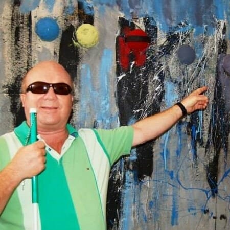 Man in dark glasses and a green striped shirt points to a large abstract painting behind him
