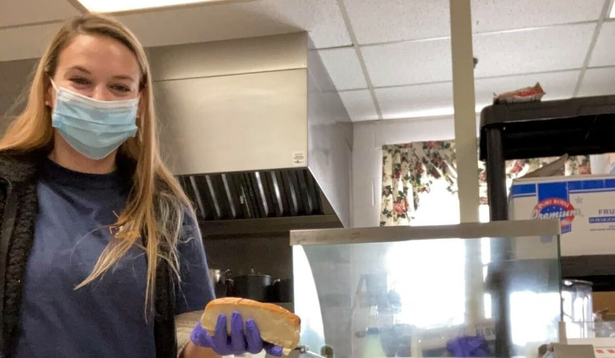 Woman dressed in blue stands holding a hotdog bun in a kitchen.