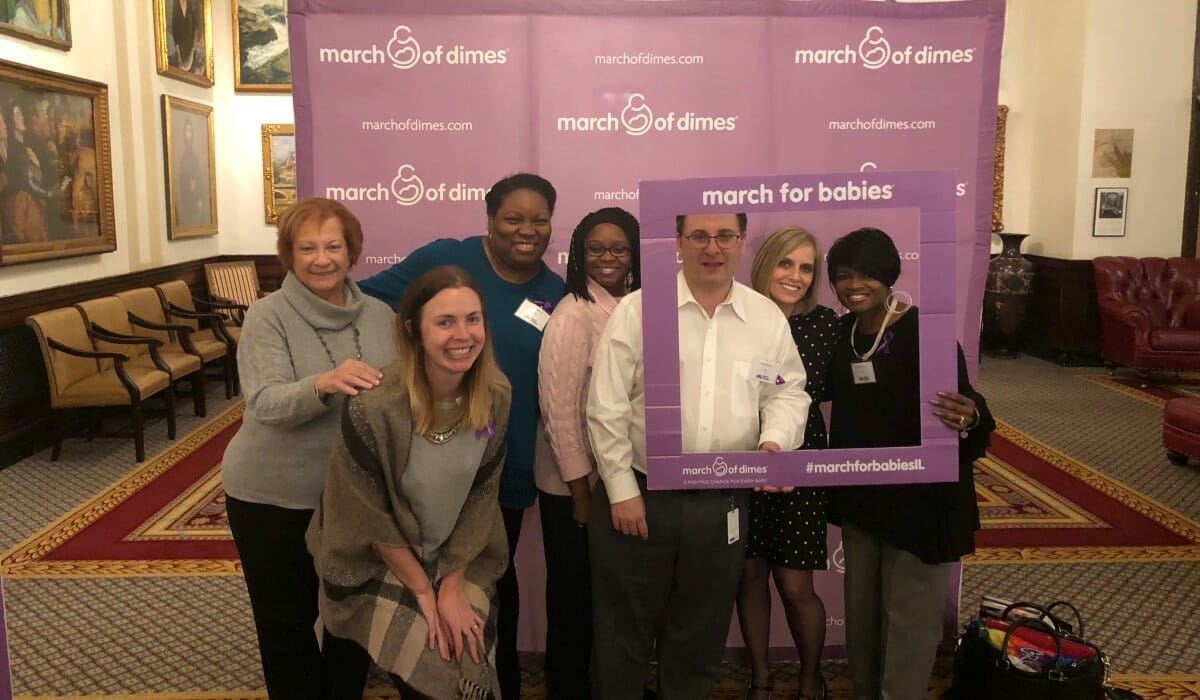 Group of people posed in front of a pink step and repeat banner emblazoned with the March of Dimes logo.