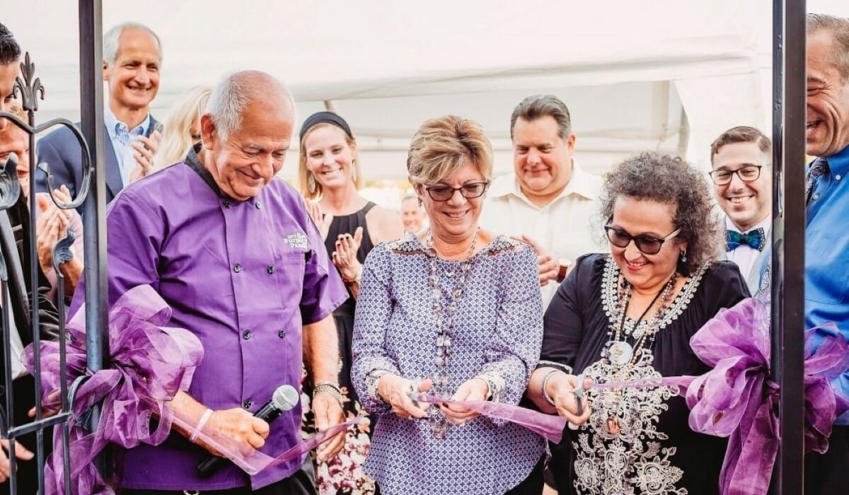 A group of smiling people, with a woman in the center cutting a purple ribbon.