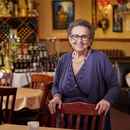 Woman with short gray hair in a purple dress stands smiling in an empty restaurant.
