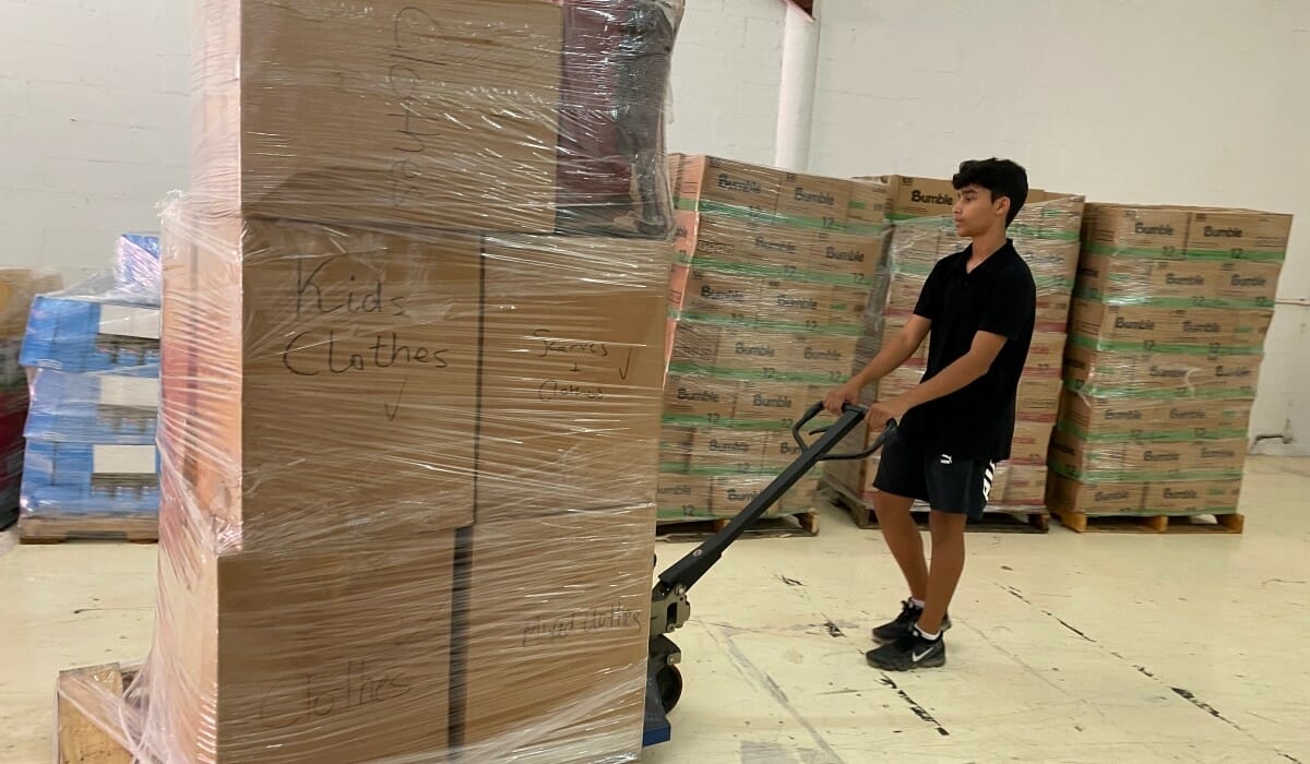 Teenage boy dressed in black pulls a pallet loaded with boxes of donated clothes in a warehouse.