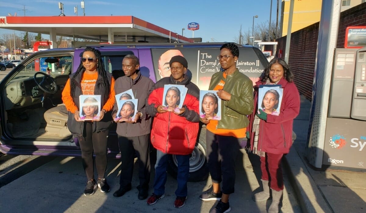 A group of people dressed in warm clothing stand outside a gas station holding photos of a missing girl. 