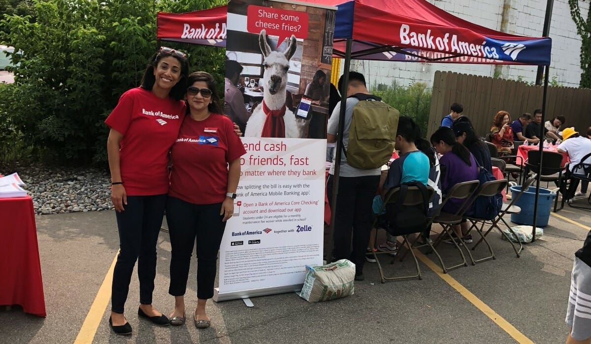 Two women in red shirts standing in front of a Bank of America tent.