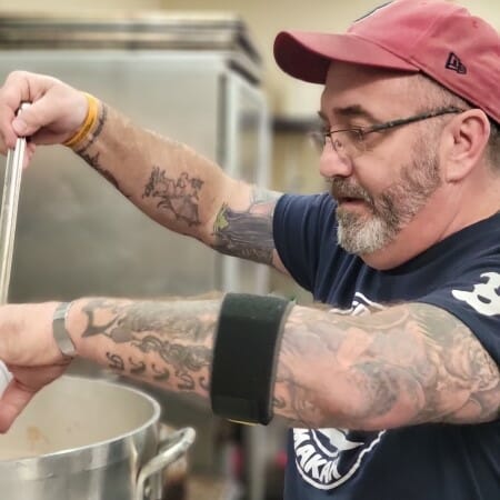 Man with glasses and tattooed arms ladles soup out of a large pot.