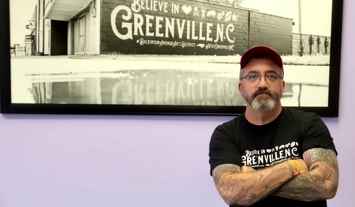 Man with glasses and tattooed arms stands in front of a photo of a building with the words Believe in Greenville, N.C.
