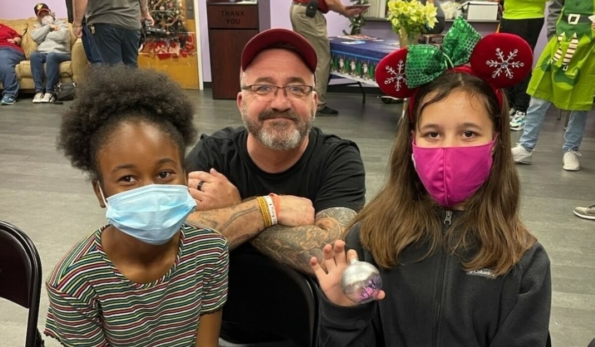 Three people pose together smiling in a room decorated for Christmas, two of them wearing masks for Covid-19 protection.