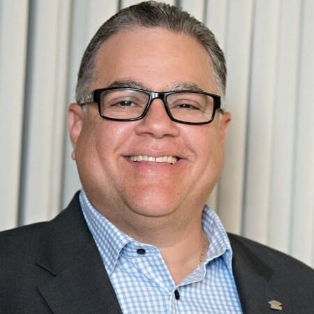 Headshot of a man in a blue checked shirt, blazer and glasses.