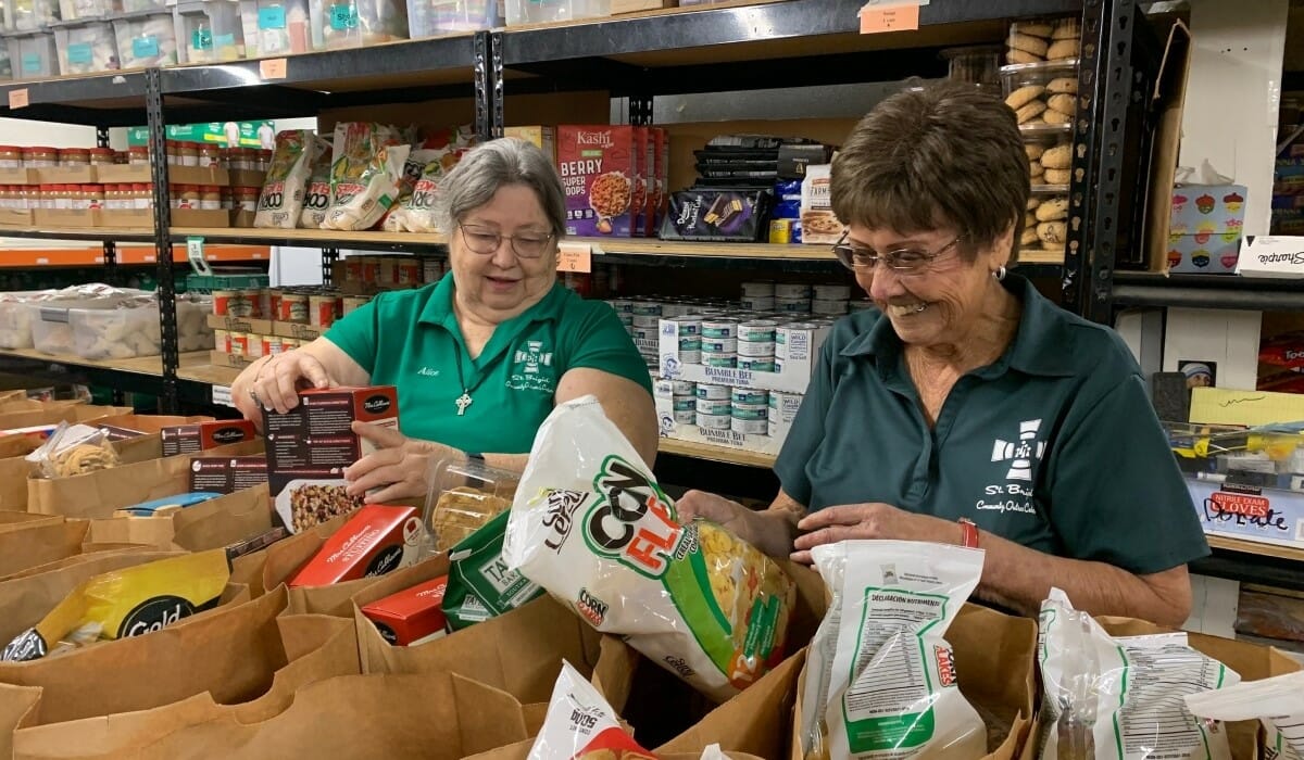 Two women in green shirts pack paper bags with donated food.