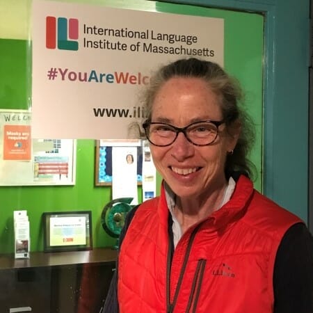Smiling woman with glasses wearing a red vest stands in front of sign reading International Language Institute of Massachussets.