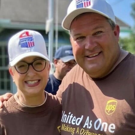 Two people in matching t-shirts and hats smile together outdoors.