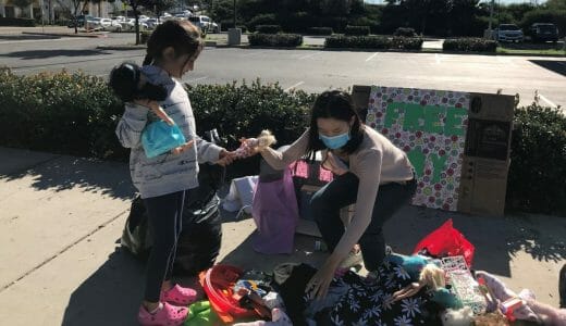 Teenage girl crouches down to hand some toys to another girl from a large pile of toys.