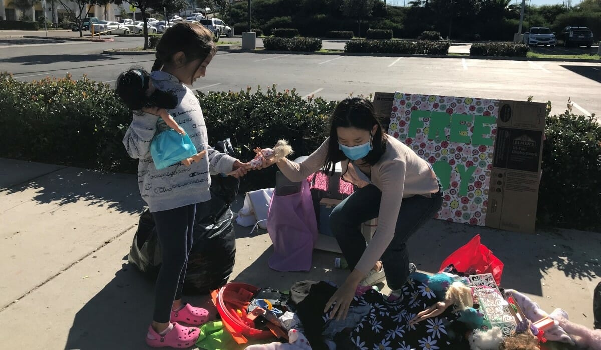 Teenage girl crouches down to hand some toys to another girl from a large pile of toys.