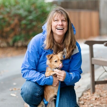 Woman in a blue rain jacket kneels on the ground holding a small brown dog.