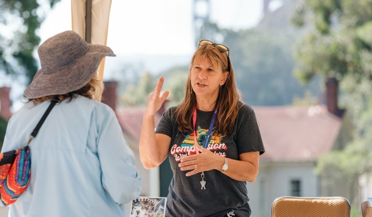Woman wearing a shirt emblazoned with a logo of the colorful image of a dog's head and the words "Compassion Without Borders" speaks animatedly.