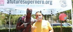 Man in a yellow shirt and man dressed in a superhero costume pose in front of a marquee tent.