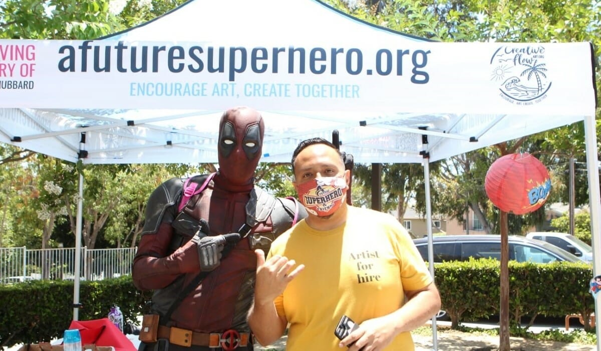Man in a yellow shirt and man dressed in a superhero costume pose in front of a marquee tent.