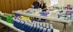 Two people stand in a room with large folding tables with essential items like shampoo and diapers.