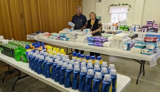 Two people stand in a room with large folding tables with essential items like shampoo and diapers.