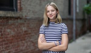 Girl in a striped shirt stands outdoors with arms folded.