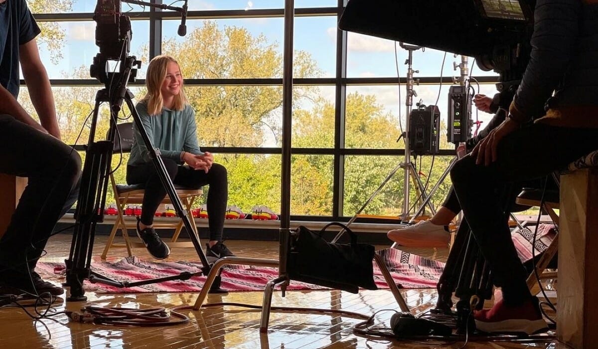 Girl in a teal hoodie sits in front of cameras for an interview.