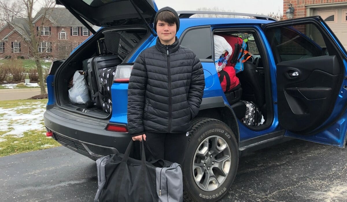Teenager in a dark parka stands in front of a car loaded with duffel bags and luggage to be donated.