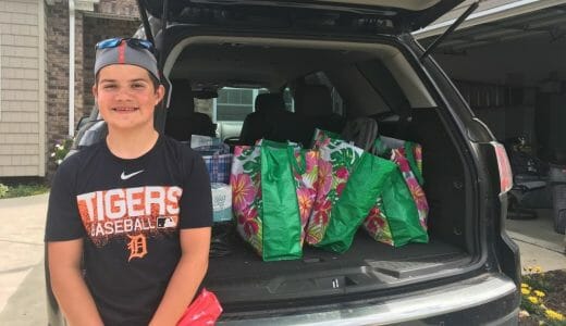 Teenager sits on the open trunk of a car loaded with bags of items to be donated.
