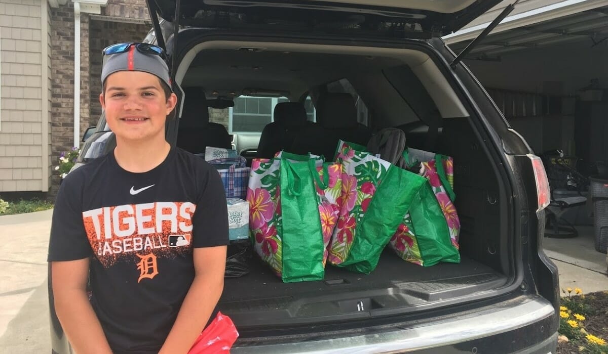 Teenager sits on the open trunk of a car loaded with bags of items to be donated.