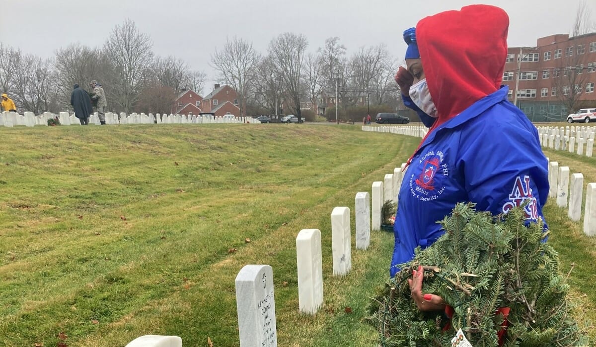 Woman holding wreathes salutes in front of a gravestone.