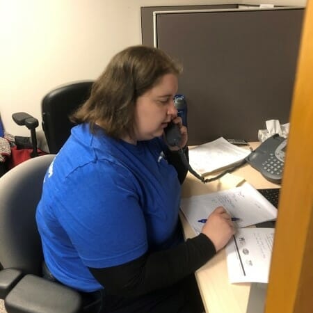 Woman in a blue shirt seated at a desk speaking on the phone.
