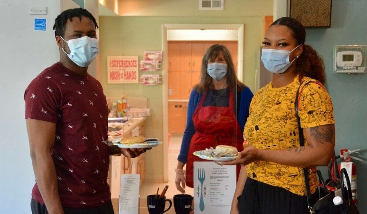 Three people in masks standing by a table with breakfast food.