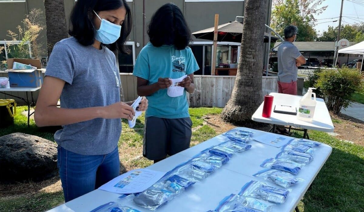 Two people in surgical masks stand behind a folding table outdoors.