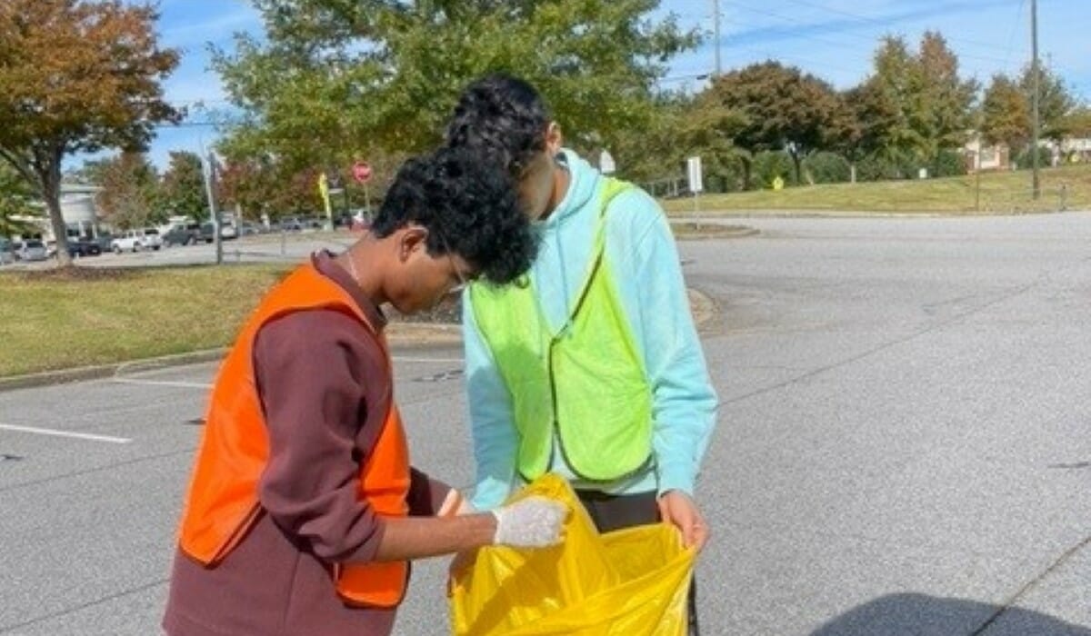 Two people in reflective vests placing trash in a large plastic bag.