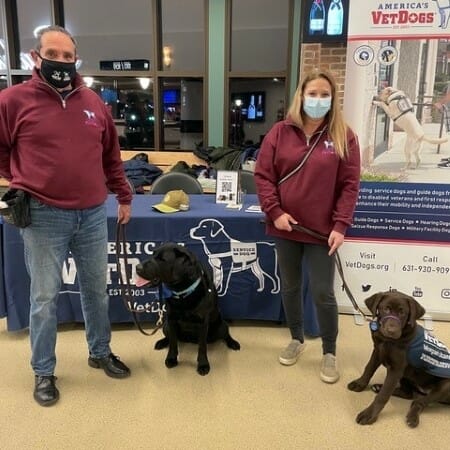 Two people in red jackets stand holding black Labradors on leashes. 