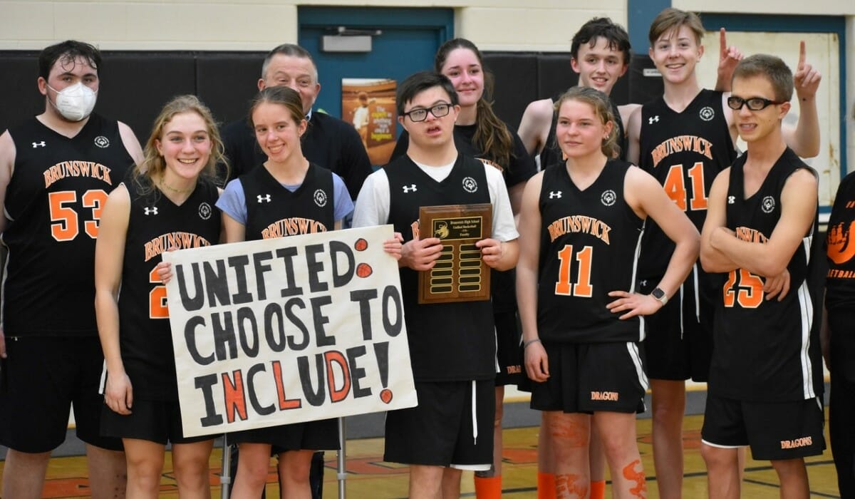 Group picture of a youth basketball team in orange and black uniforms. One player holds a sign reading "Unified: Choose to Include!"