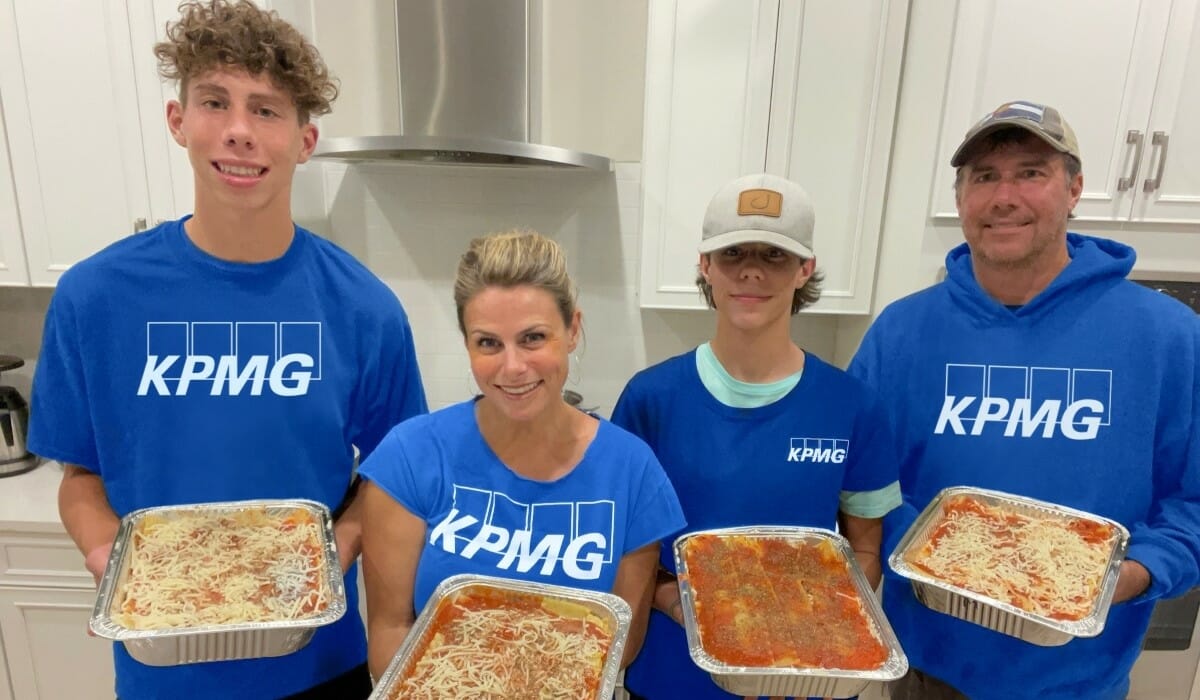 Four people wearing matching blue shirts emblazoned with the letters "KPMG" pose holding pans of lasagna.