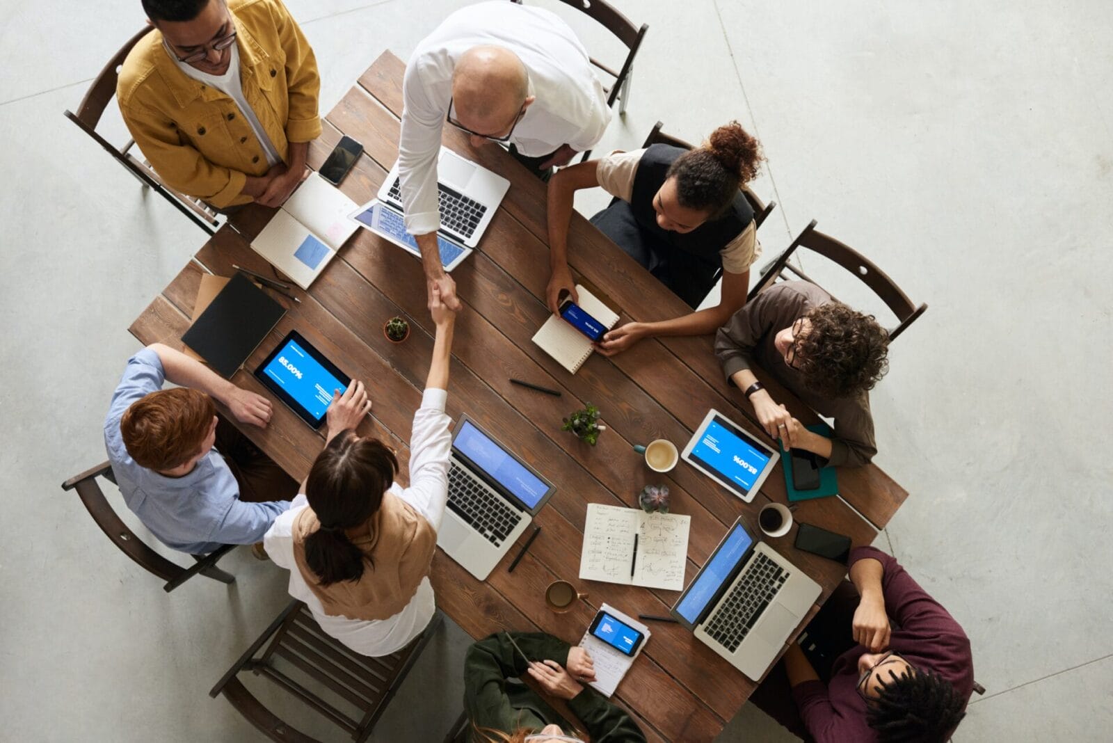 Aerial view of people sitting at a conference table with laptops. Two people are shaking hands across the table.