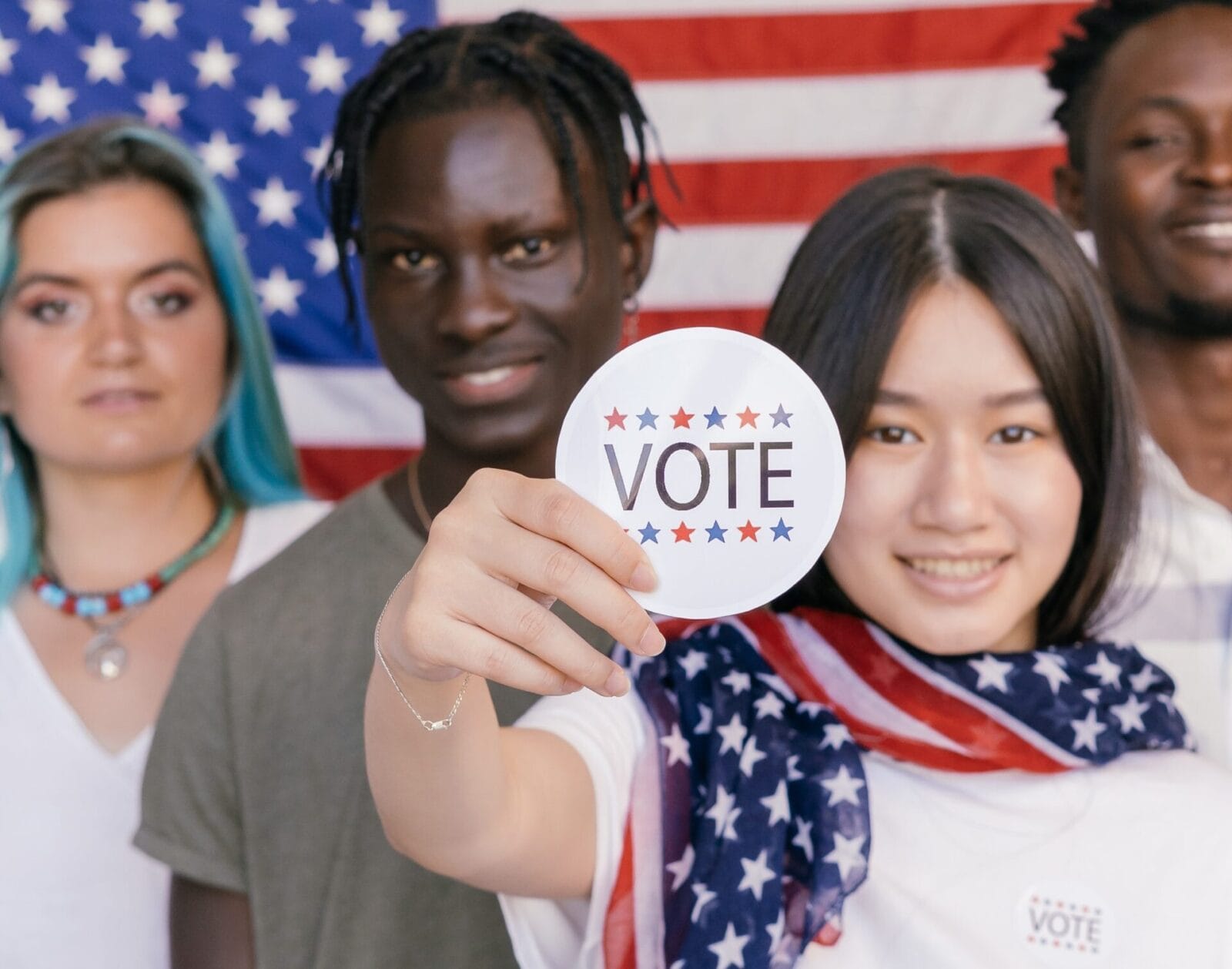 Multi-cultural group of four young people standing in front of an American flag. The female-presenting person is holding up a "vote" sticker.