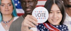 Multi-cultural group of four young people standing in front of an American flag. The female-presenting person is holding up a "vote" sticker.