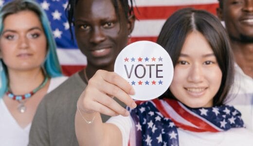 Multi-cultural group of four young people standing in front of an American flag. The female-presenting person is holding up a "vote" sticker.